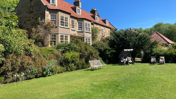 A large stone building with multiple windows and a red-tiled roof, surrounded by lush greenery and a grassy lawn.