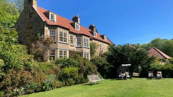 A large stone building with multiple windows and a red-tiled roof, surrounded by lush greenery and a grassy lawn.