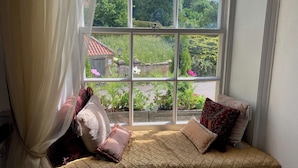 A window seat with cushions and a view of a garden and a building with a red roof.