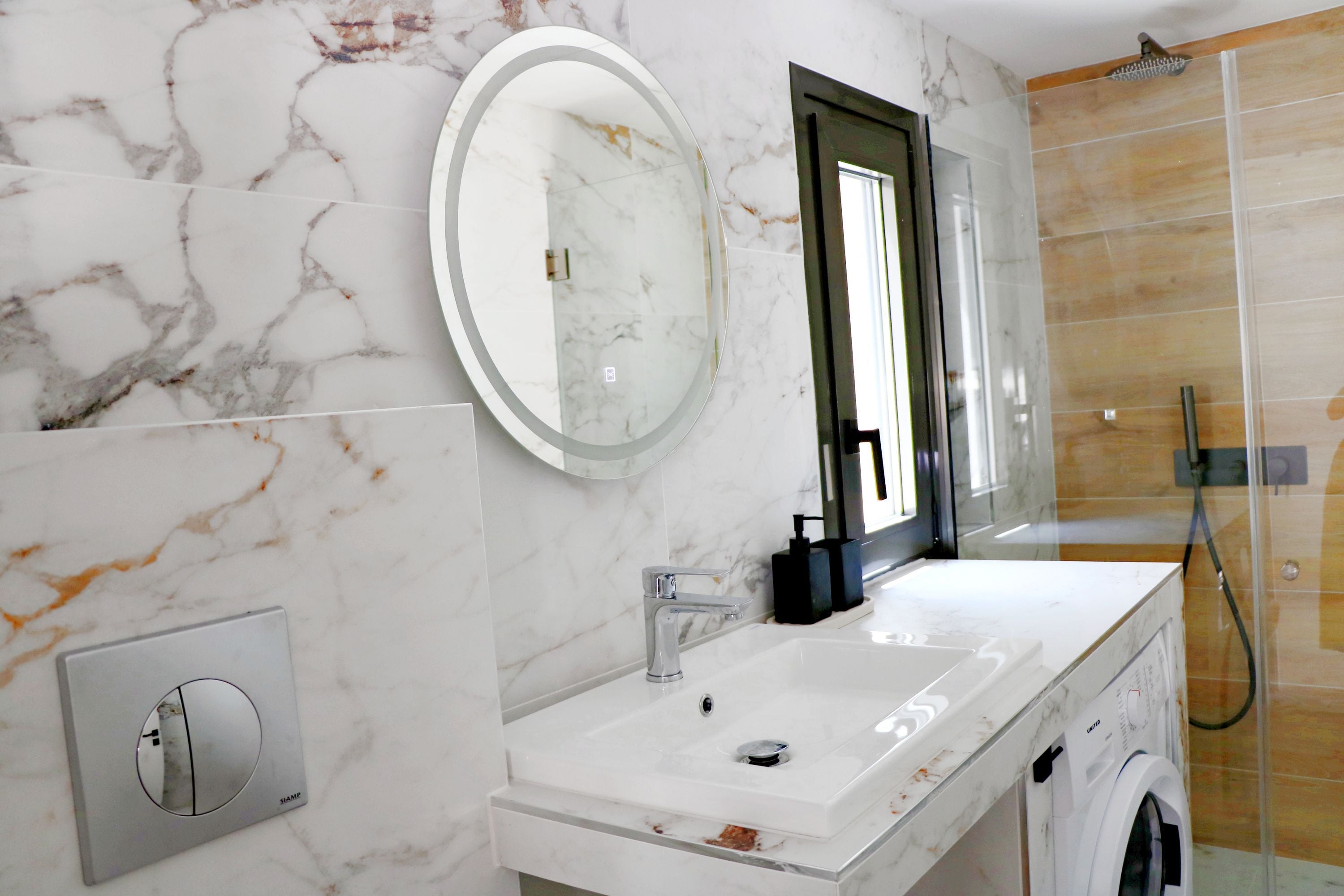 Un baño con pared de mármol, un espejo redondo, un lavamanos, un grifo, una ducha y una lavadora.