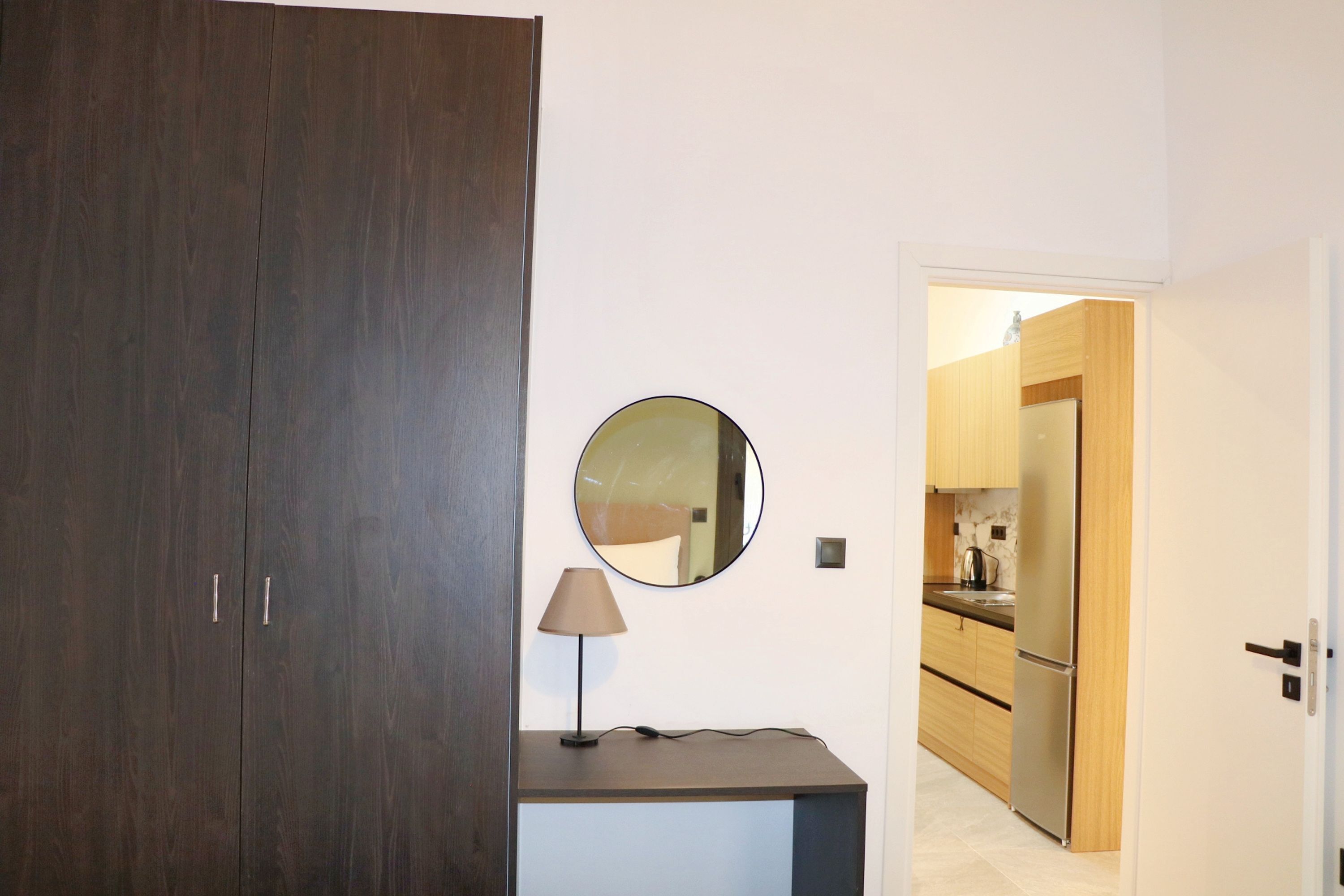 A room with a dark wooden wardrobe, a small table with a lamp, a round mirror, and a view into a kitchen area with wooden cabinets and a refrigerator.