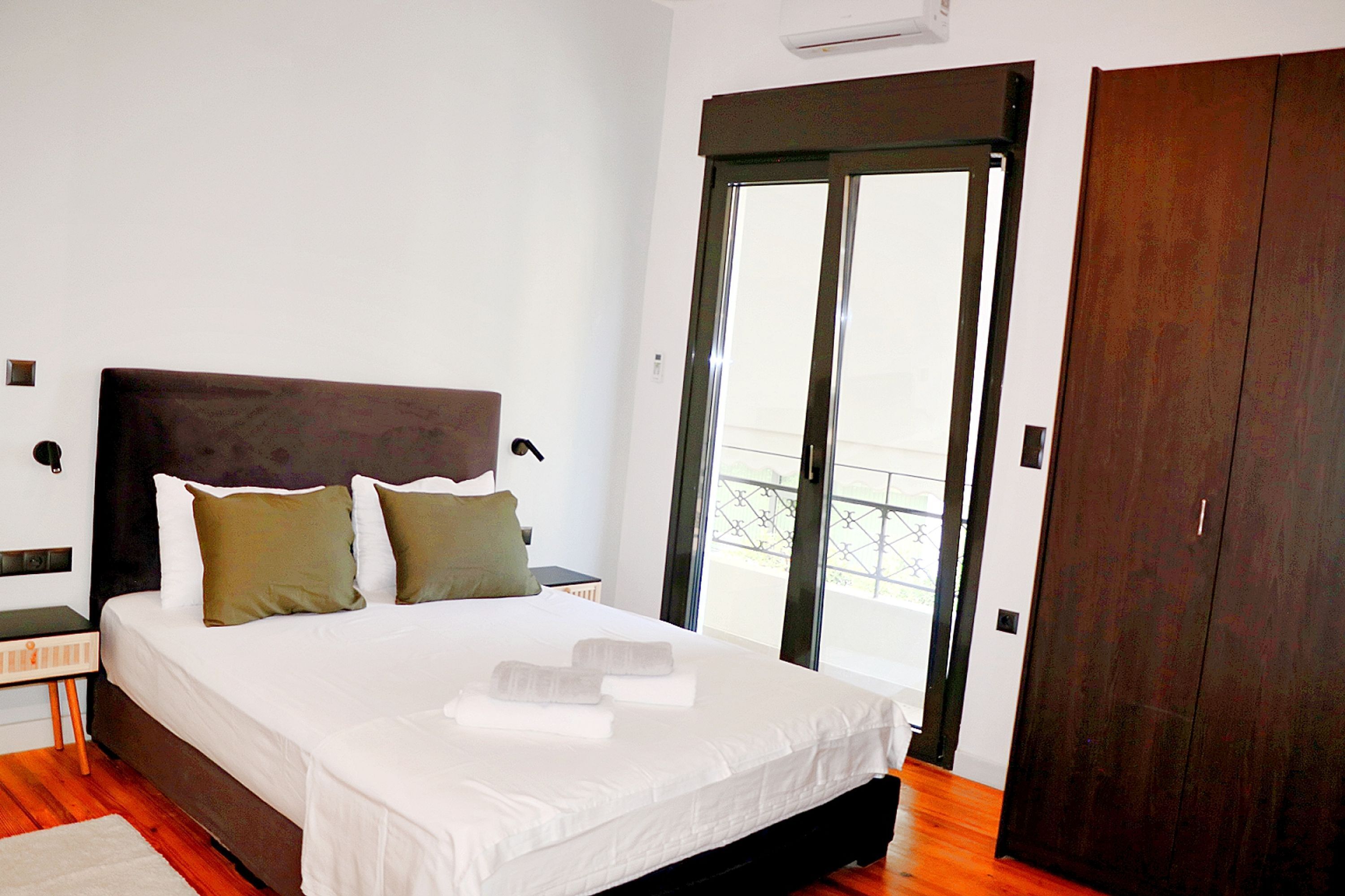 A bedroom with a large bed, a wooden wardrobe, and a sliding glass door to the balcony.