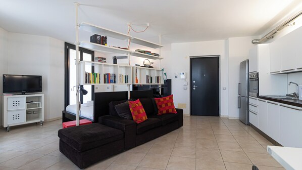 A modern living room with a black sofa, red cushions, a flat-screen TV, a bookshelf, and a kitchen area with white cabinets and a black countertop. - Italianway Easy - Segesta 3 (San Siro)