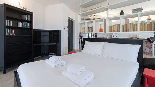 A hotel room with a large bed, a bookshelf, a desk with a computer, and a small kitchen area. - Italianway Easy - Segesta 3 (San Siro)