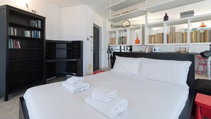 Ein Hotelzimmer mit einem großen Bett, einer Bücherwand, einem Schreibtisch mit Computer und einer kleinen Küchenzeile.