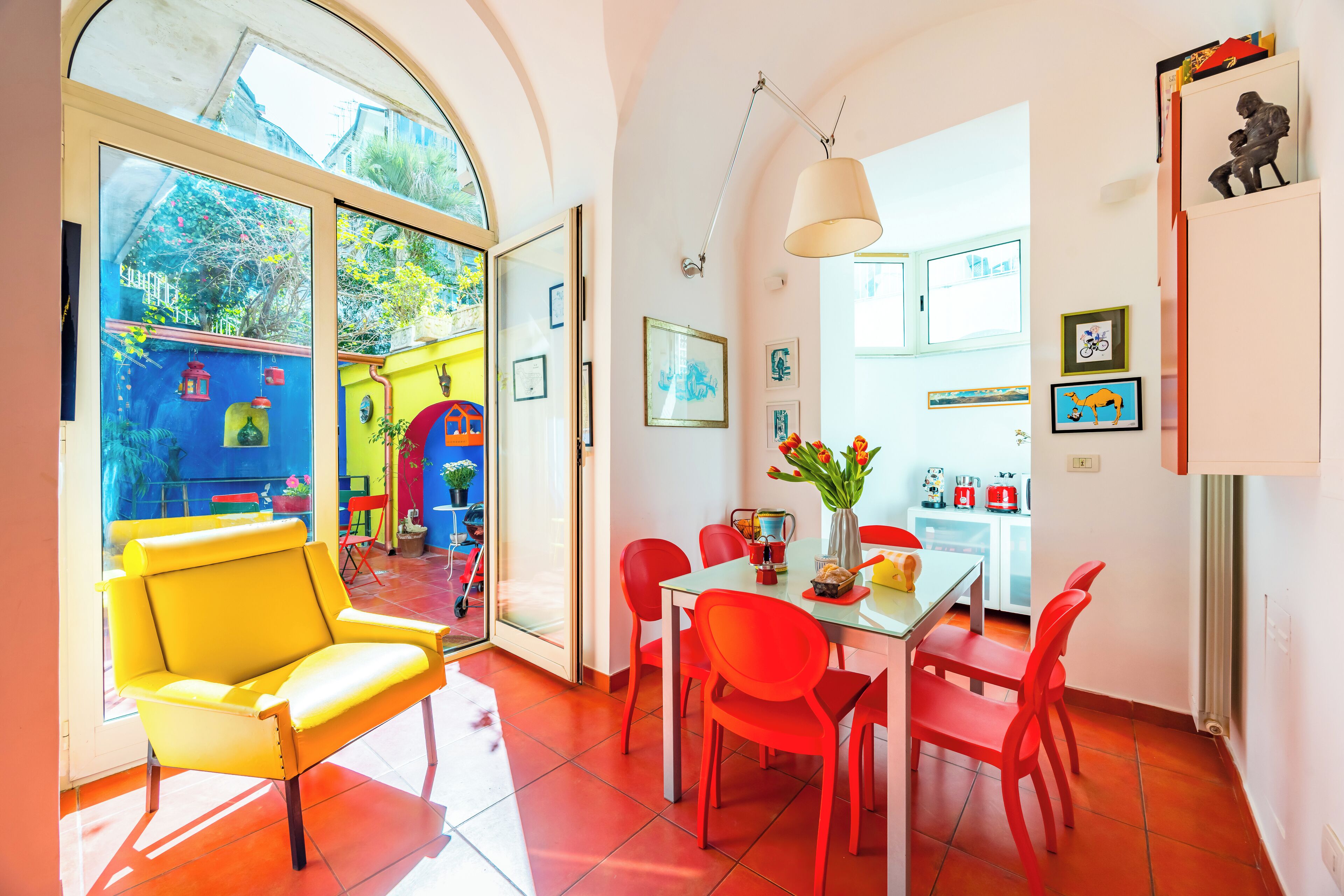 Ein heller Essbereich mit Tisch und Stühlen, ein gelber Sessel und ein großes Fenster mit Blick auf einen farbenfrohen Außenbereich.