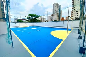 Una cancha de baloncesto interior con suelo azul y amarillo, rodeada por una pared blanca y vallas metálicas.
