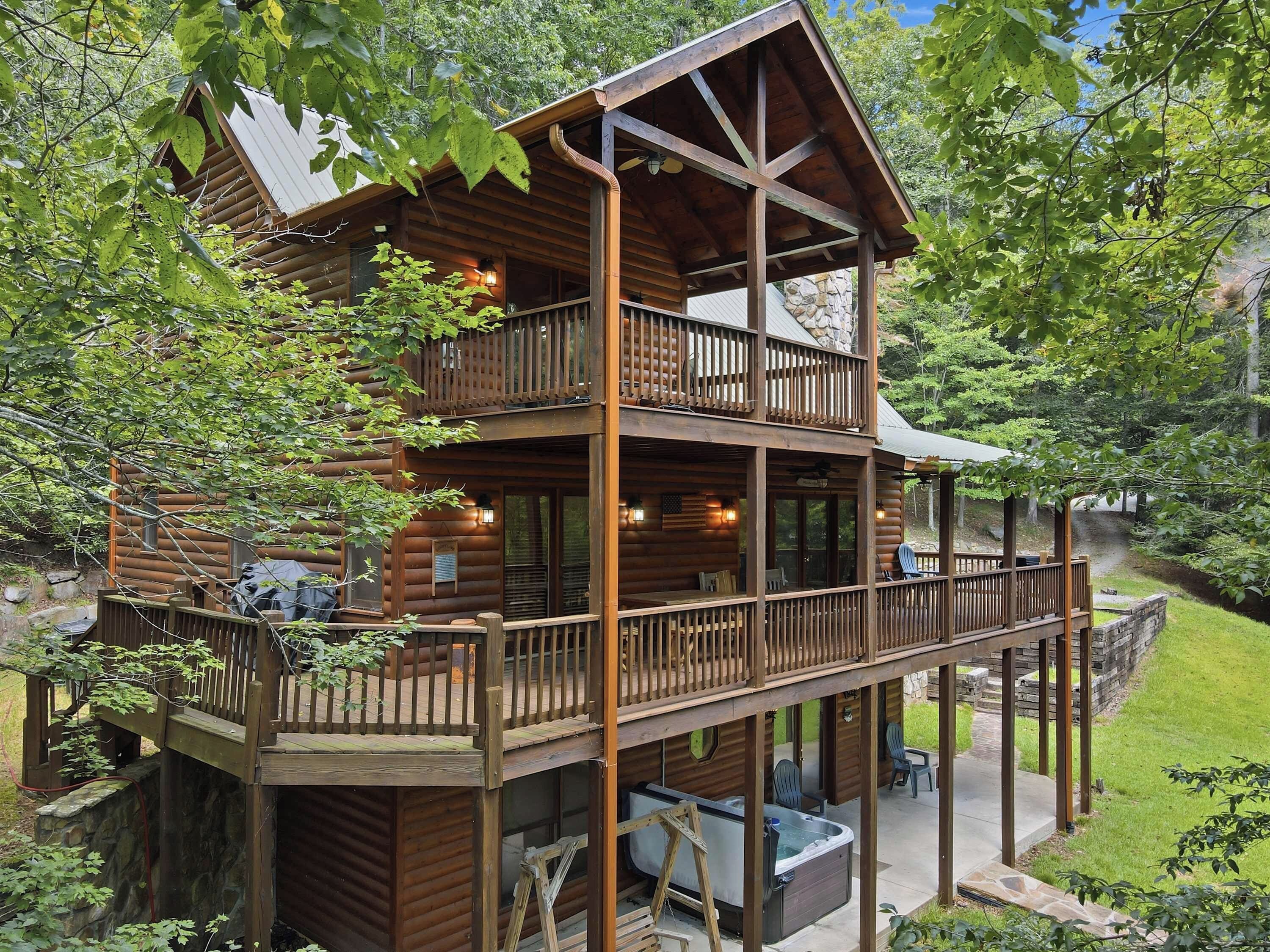 Une cabane en rondins à deux étages, dotée d’un balcon, d’un jacuzzi et d’un espace extérieur aménagé, le tout entouré d’arbres.