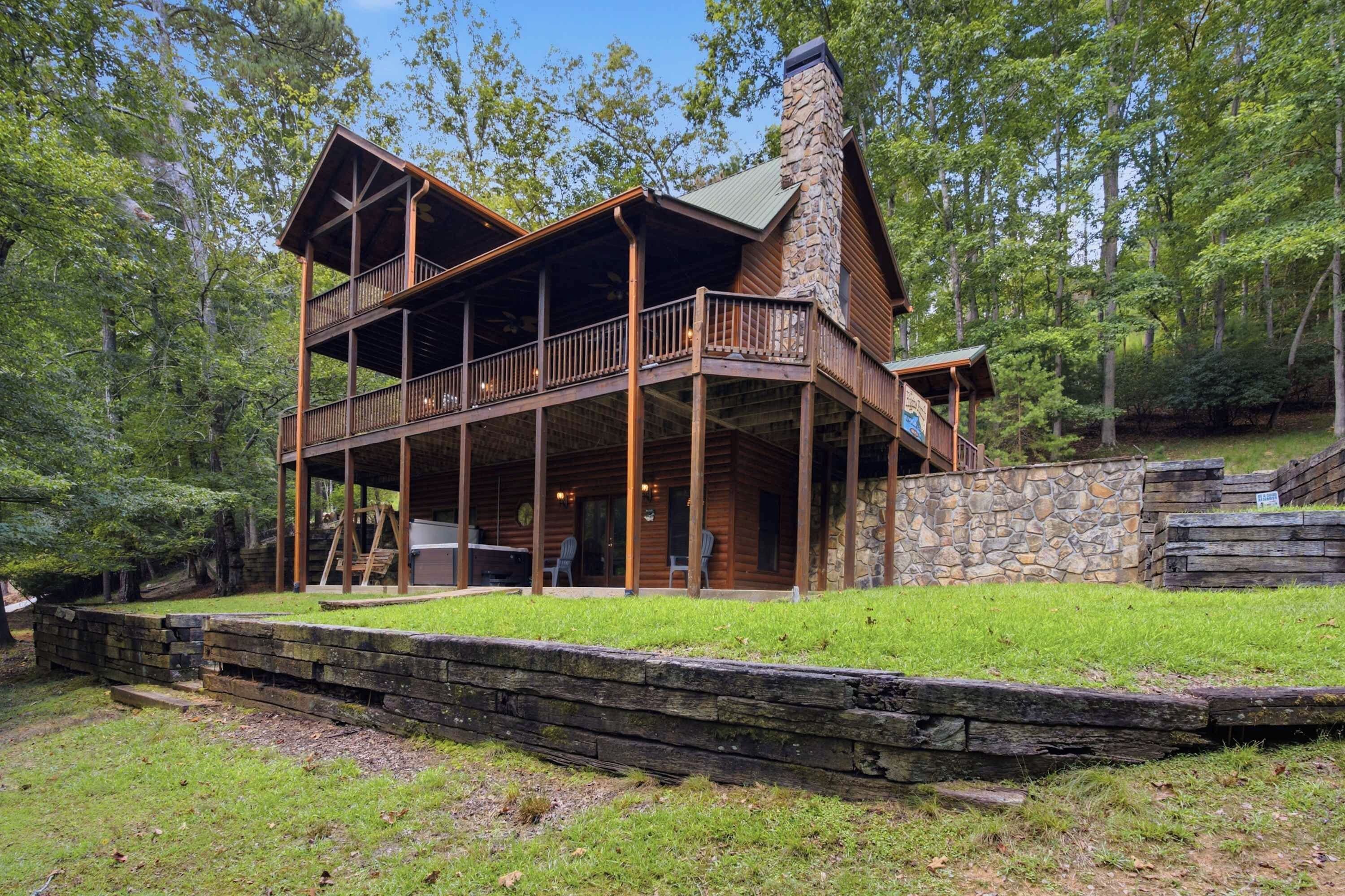 A two-story log cabin with a stone chimney, a covered porch, and a stone retaining wall.