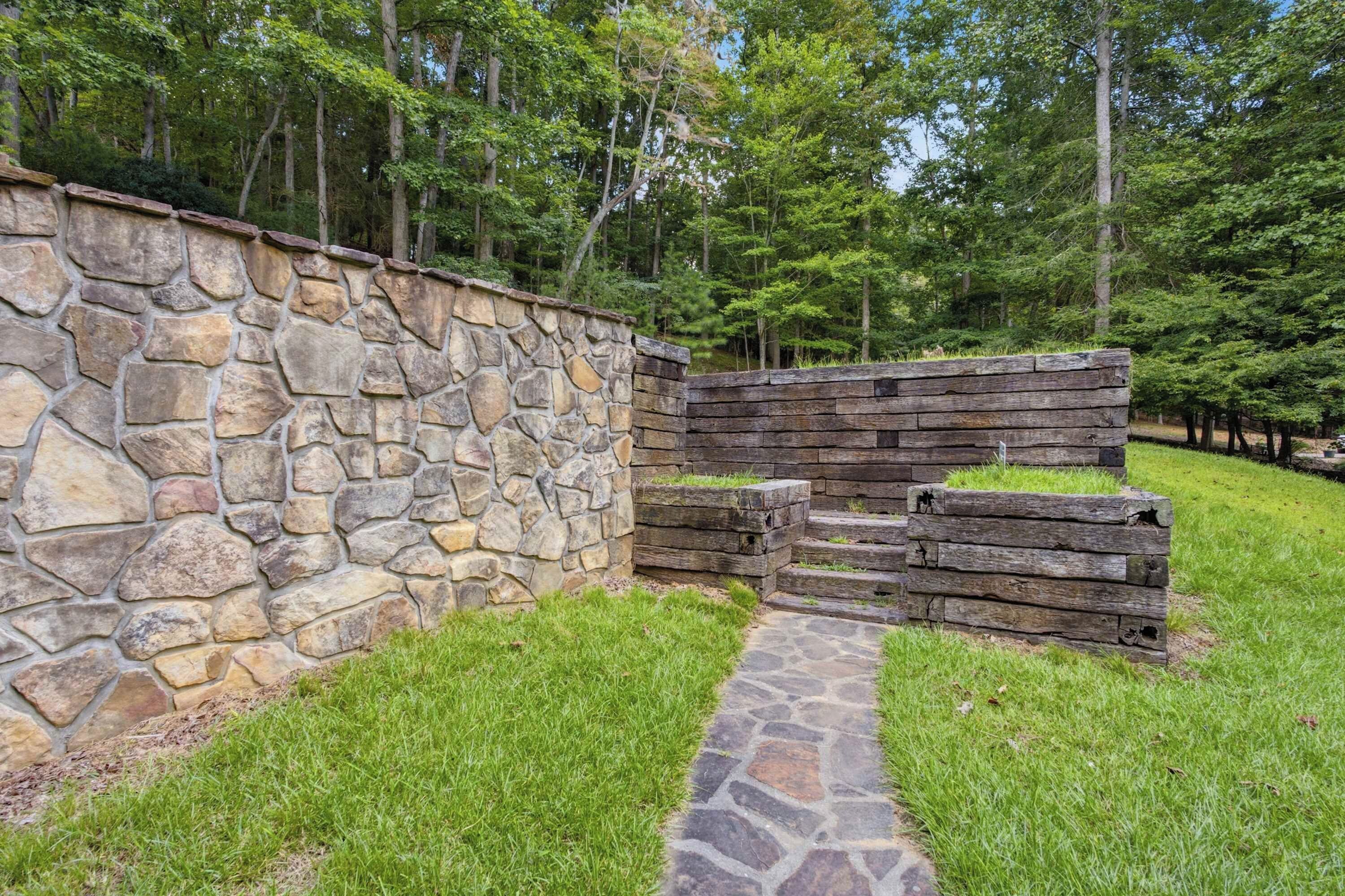 A stone wall with a wooden structure adjacent to it, a stone pathway leading through a grassy area, and a forested background.