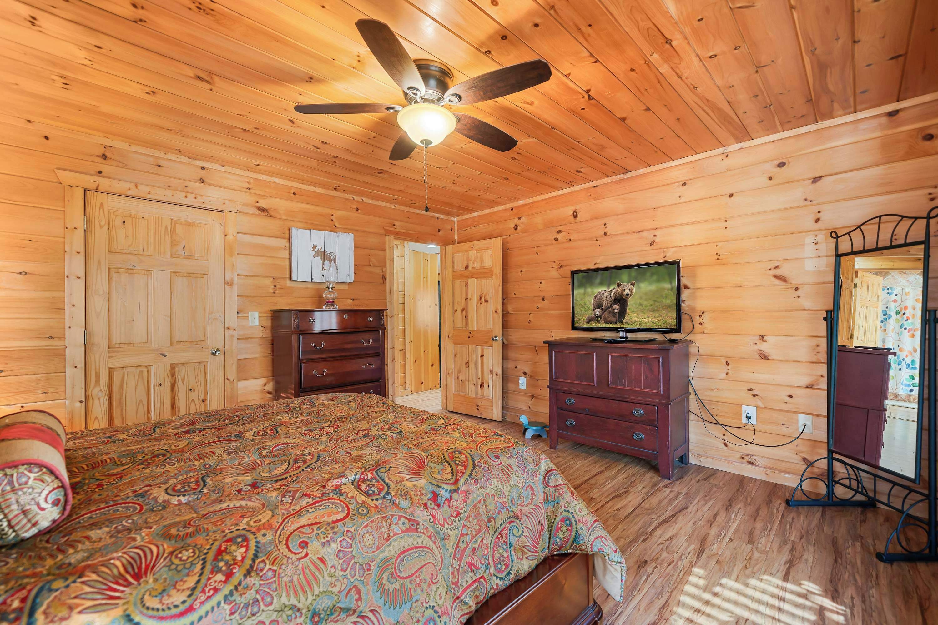 Een slaapkamer in een houten chalet met een bed, een dressoir, een televisie en een plafondventilator.