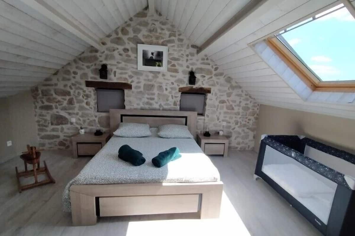 A bedroom with a stone wall, a bed with pillows, a nightstand, a wooden chair, and a skylight.