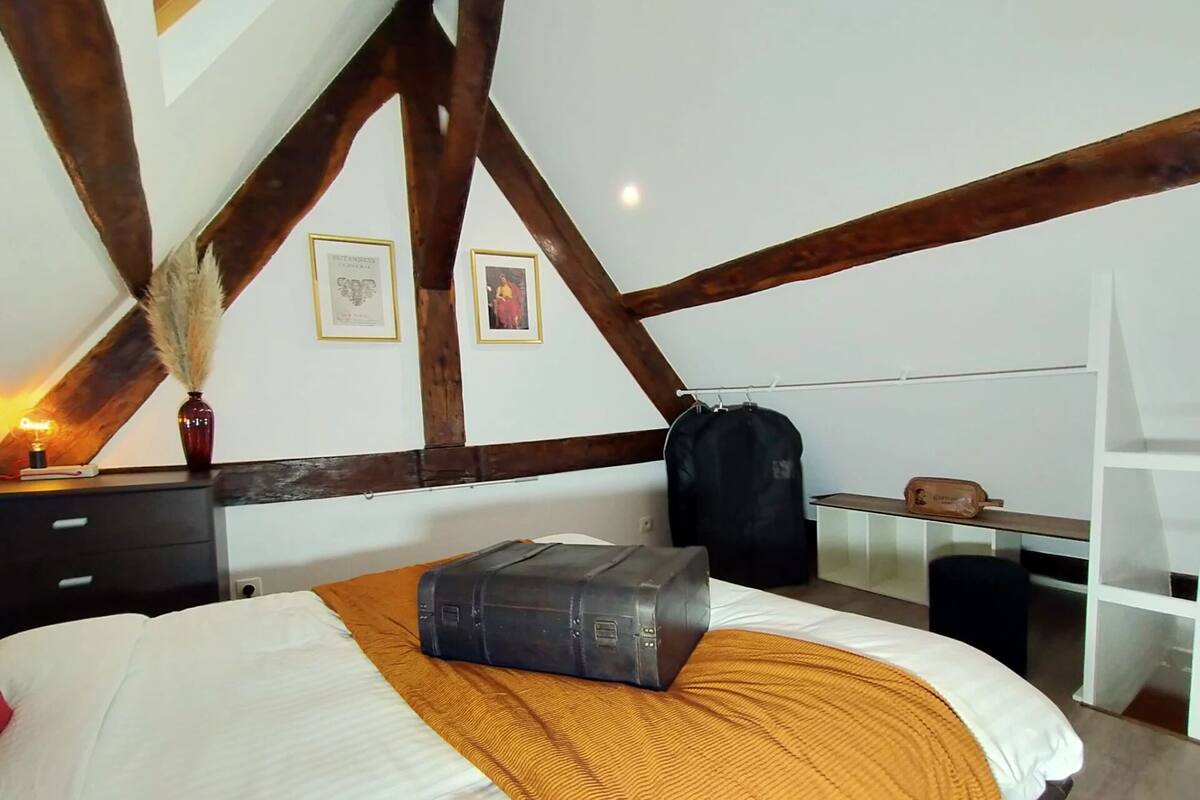 A bedroom with a high ceiling, exposed wooden beams, a bed with a white and orange bedspread, a suitcase on the bed, a desk with a lamp, and framed pictures on the wall.