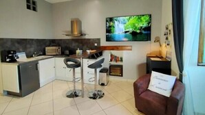 A modern kitchen with a microwave, toaster, and coffee machine. There is a TV mounted on the wall displaying a nature scene. A bar stool is in the center, and a red chair with a pillow is on the right. - Manoir de l'Aisnerie (Saint-Herblain)