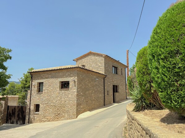 Un edificio de piedra con techo de tejas, una calle pavimentada y un gran arbusto verde.