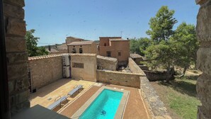 Zona de piscina con paredes de piedra, sillas de descanso, un edificio de piedra con tejado de baldosas y cielo azul despejado.