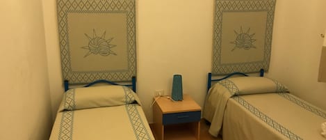 A room with two single beds, each with a patterned bedspread and matching curtains. There is a small wooden nightstand between the beds with a lamp on it.