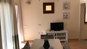 A dining area with a table, chairs, a vase, a television on a white stand, and a wall-mounted air conditioner. - Residenze del Mare (Porto Ottiolu)