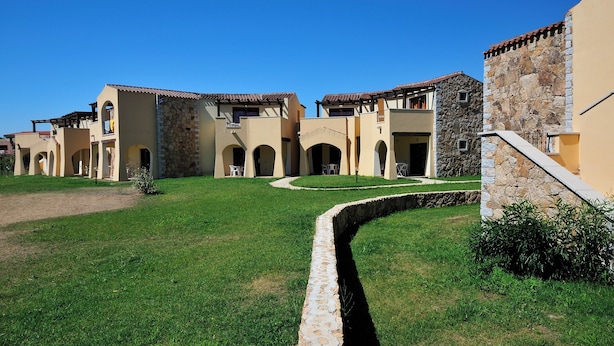 A courtyard with a stone wall, a grassy area, and buildings with arched doorways and balconies. - Residenze del Mare (Porto Ottiolu)