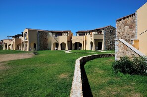 A courtyard with a stone wall, a grassy area, and buildings with arched doorways and balconies. - Residenze del Mare (Porto Ottiolu)