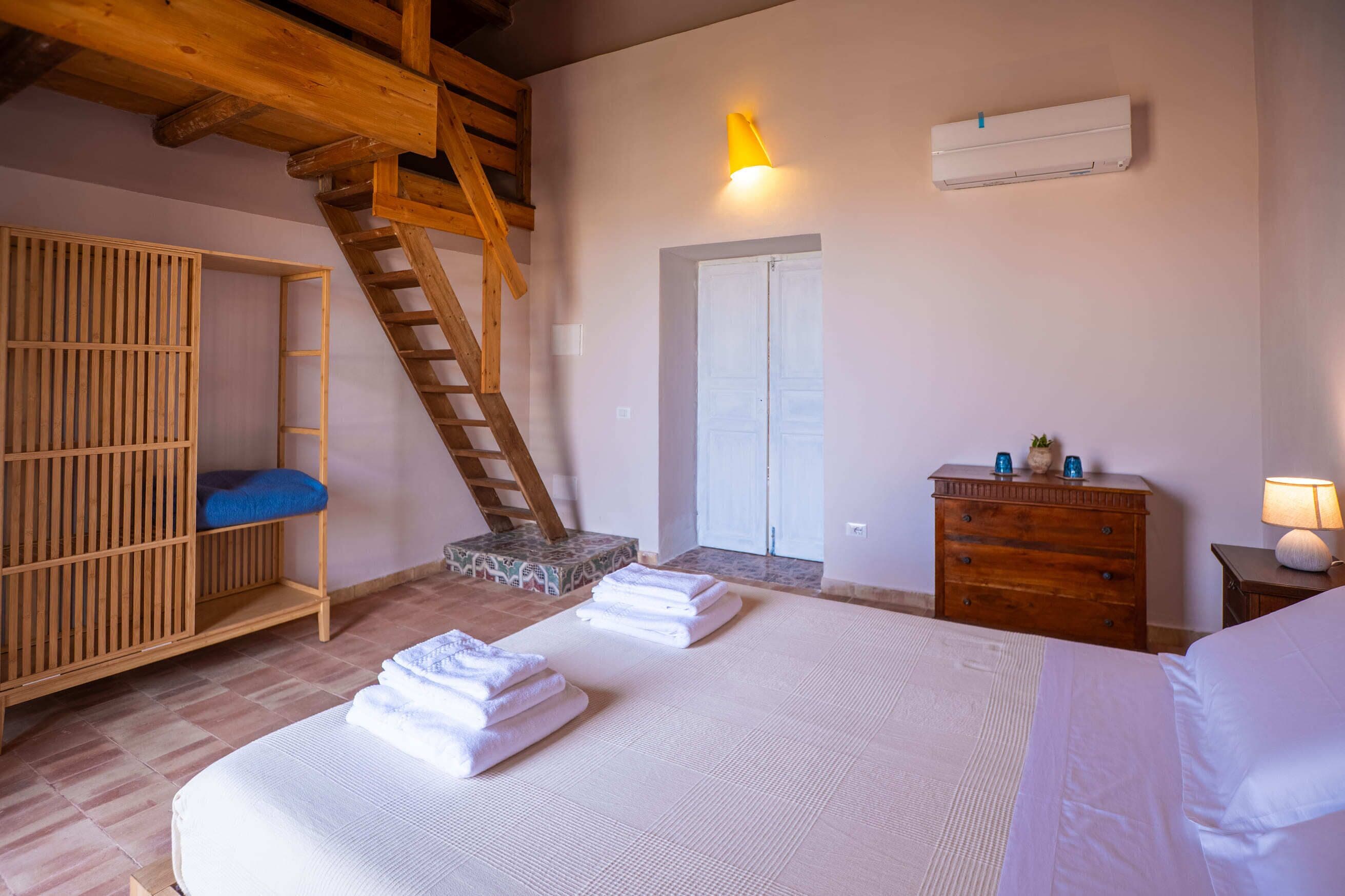 A bedroom with a bed, a wooden wardrobe, a dresser, a ladder, an air conditioning unit, and a door.