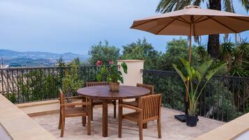 Een ronde houten tafel met stoelen en een grote parasol op een balkon met uitzicht over het landschap.