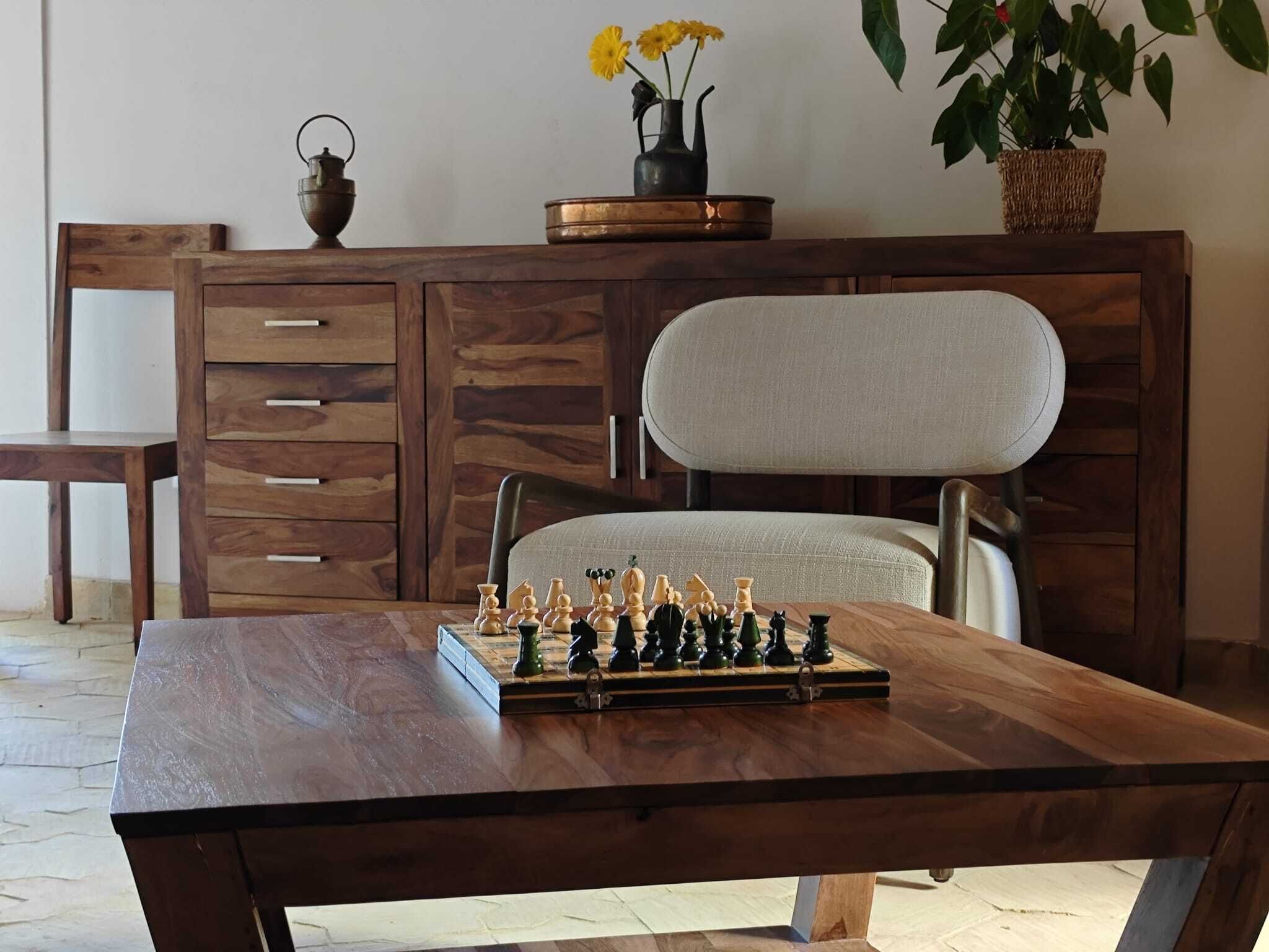 Una mesa de madera con un tablero de ajedrez, una silla, un aparador con cajones y una planta en maceta.