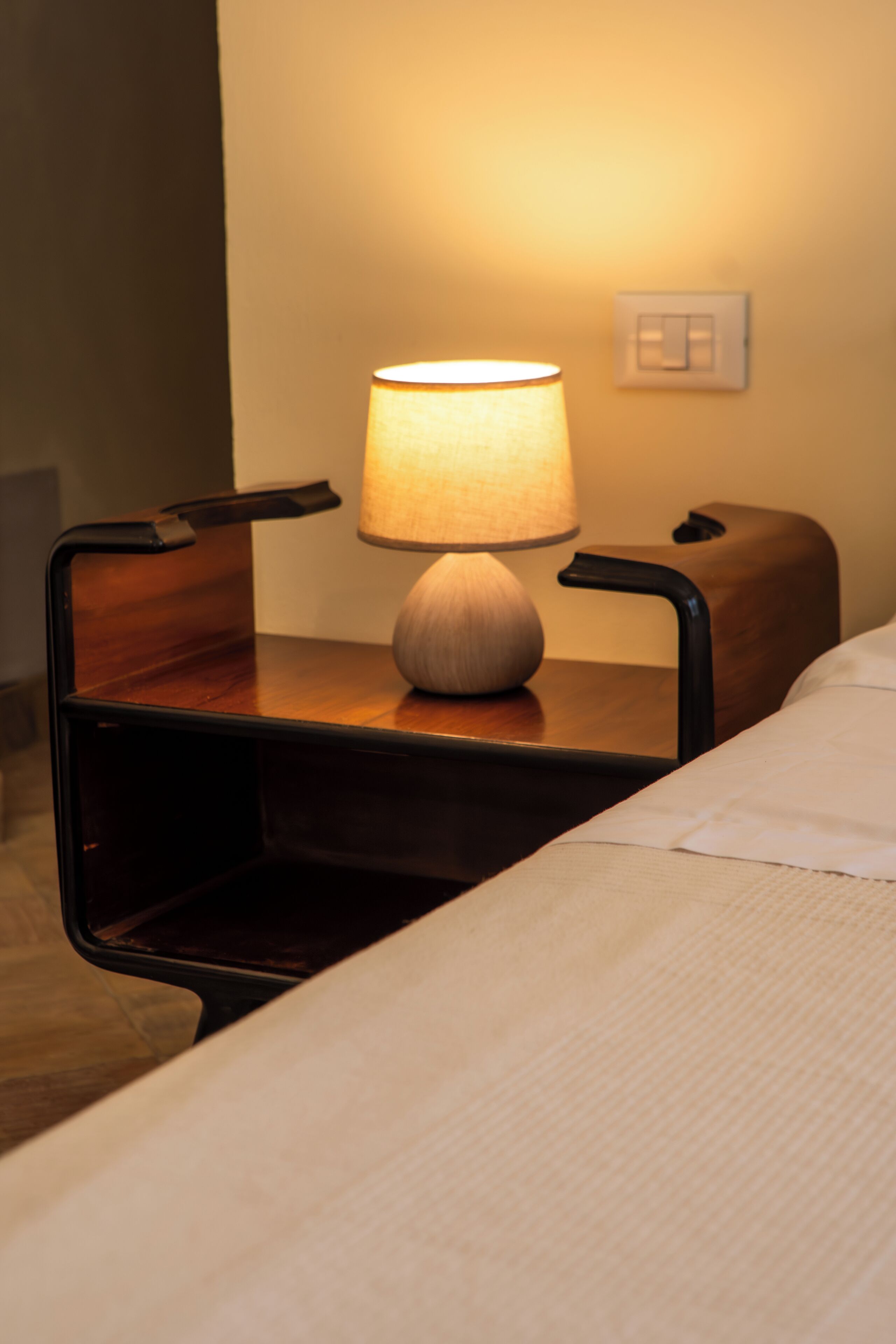 A hotel room with a wooden nightstand, a lamp, a chair, and a bed with a white sheet.