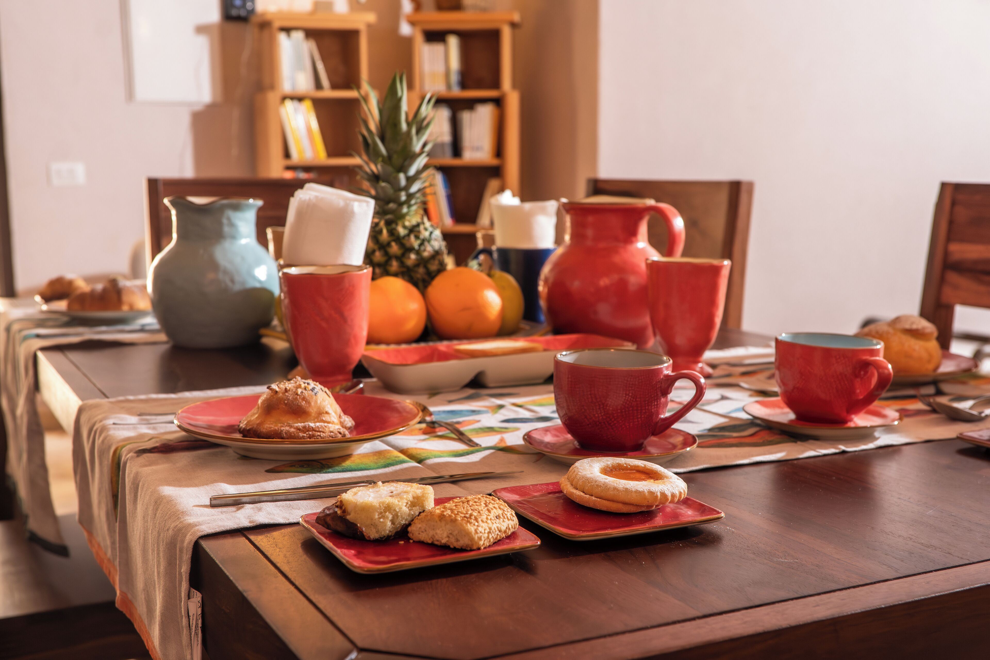A dining table set with plates, cups, and a pitcher, surrounded by bookshelves and a pineapple.