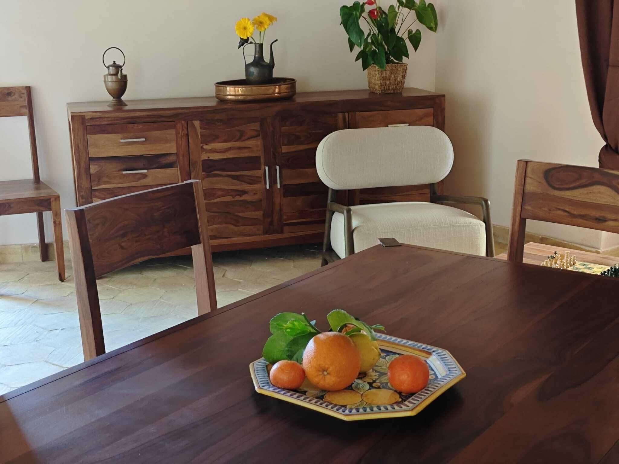 A dining area with a wooden table, chairs, a fruit plate, a vase with flowers, and a decorative item on a wooden cabinet.