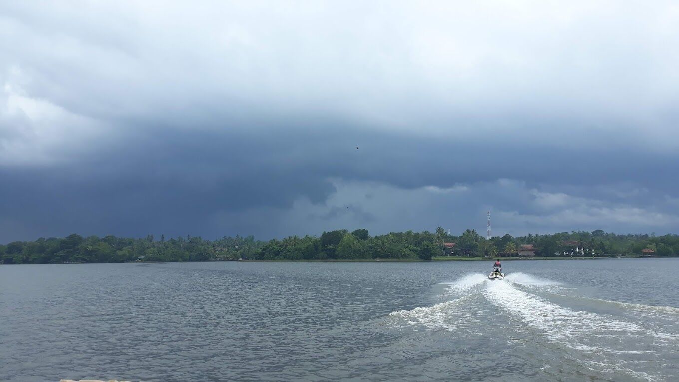 A body of water with a jet ski moving across it, a cloudy sky, and a distant shoreline with vegetation and buildings.