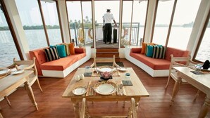 A dining area with a wooden table set for four, surrounded by rattan chairs and cushioned sofas with colorful pillows. A person is visible in the background near the railing. - Houseboats By DAE Colombo (Piliyandala)