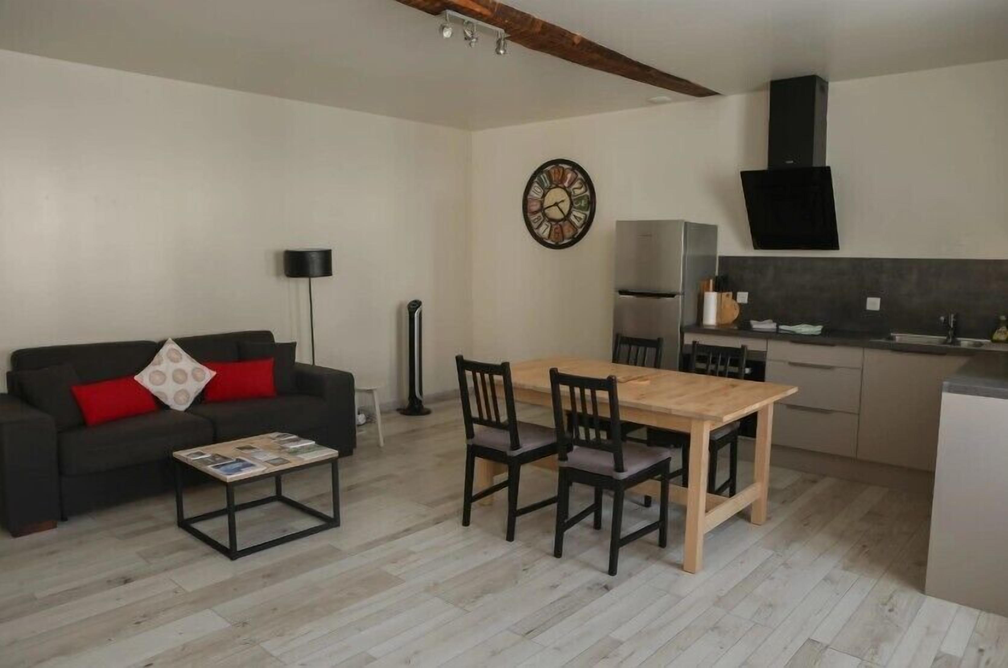 A compact living space with a sofa, a dining table with chairs, a coffee table, a clock, a modern kitchen with a stove, and a black speaker.