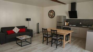 A compact living space with a sofa, a dining table with chairs, a coffee table, a clock, a modern kitchen with a stove, and a black speaker.