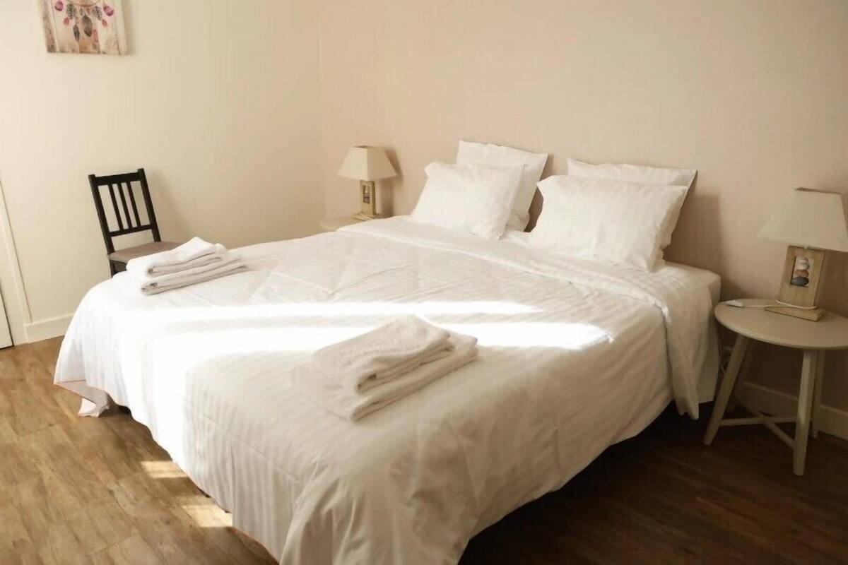 Un lit bien fait, avec du linge de lit et des serviettes blanches, une petite chaise en bois et deux tables de chevet avec des lampes.