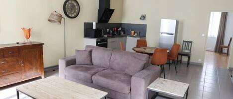 Un salon comprenant un canapé, une table basse et un coin repas avec des chaises. Au fond, on aperçoit une cuisine équipée d’une cuisinière, d’un four à micro-ondes et d’un réfrigérateur.