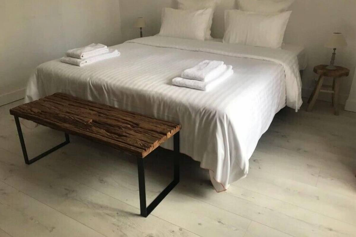 Un lit bien fait, avec du linge de lit blanc et des serviettes pliées, un banc en bois et un petit tabouret dans une pièce minimaliste au revêtement de sol de couleur claire.