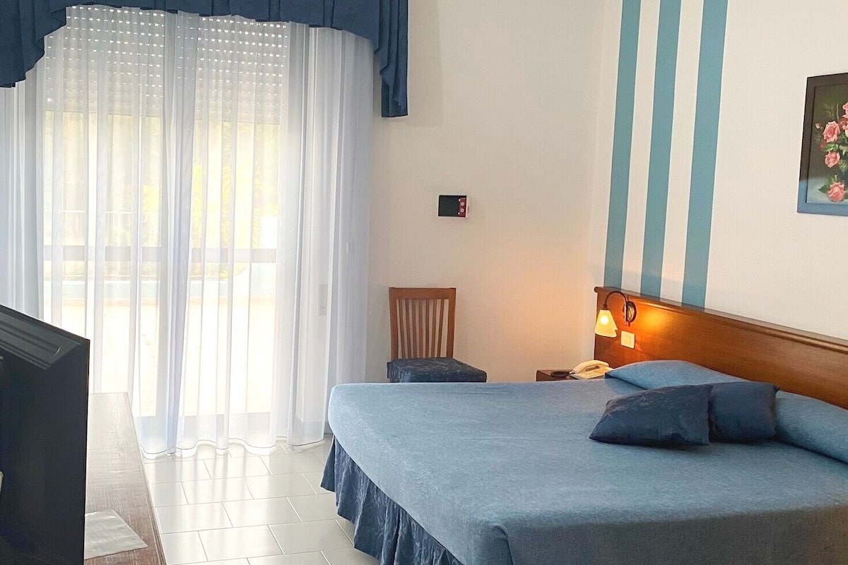 Une chambre d’hôtel avec un lit, une table de chevet, une chaise, un téléviseur et une fenêtre avec des rideaux.