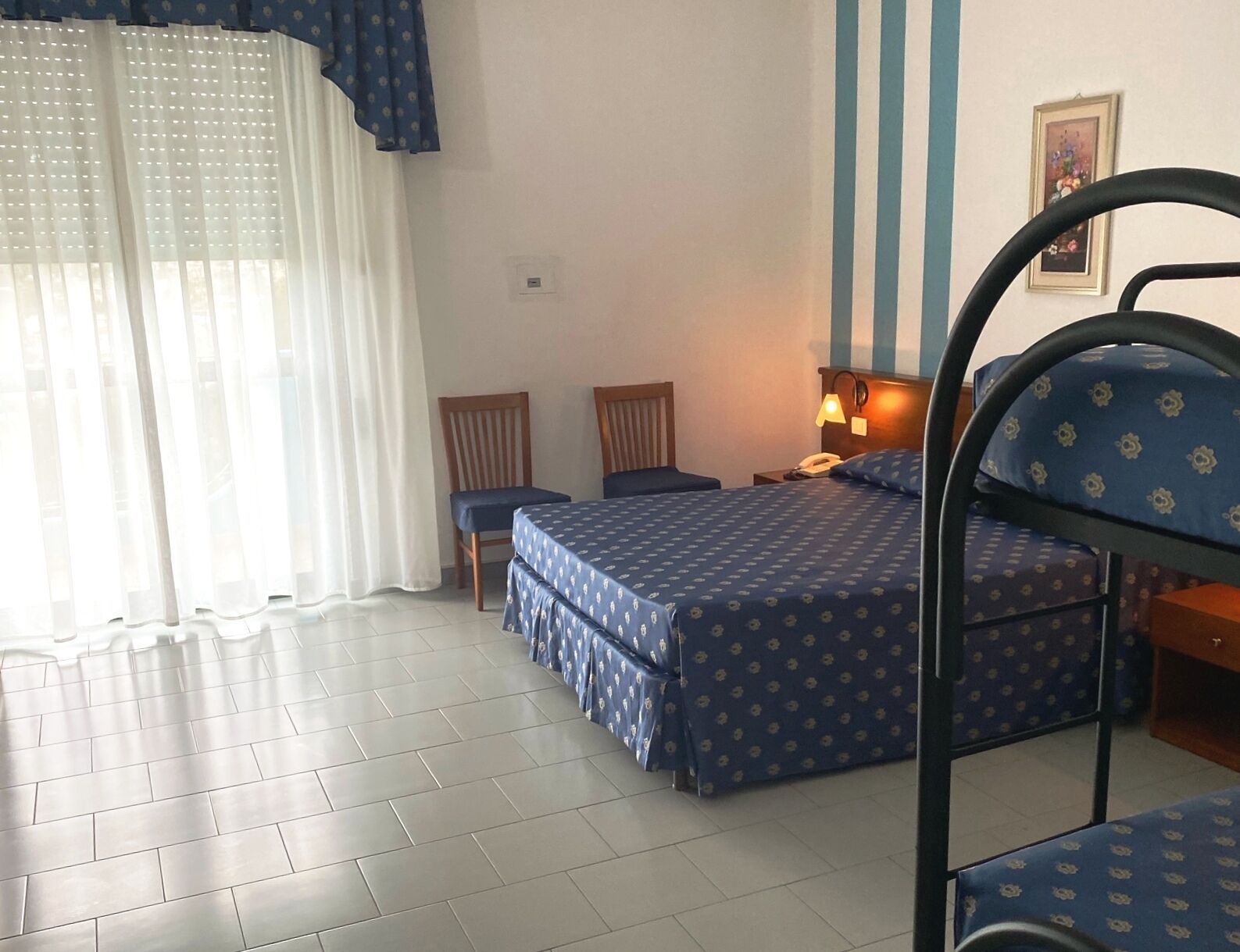 A hotel room with a bed, two chairs, a desk with a lamp, and a window with curtains.
