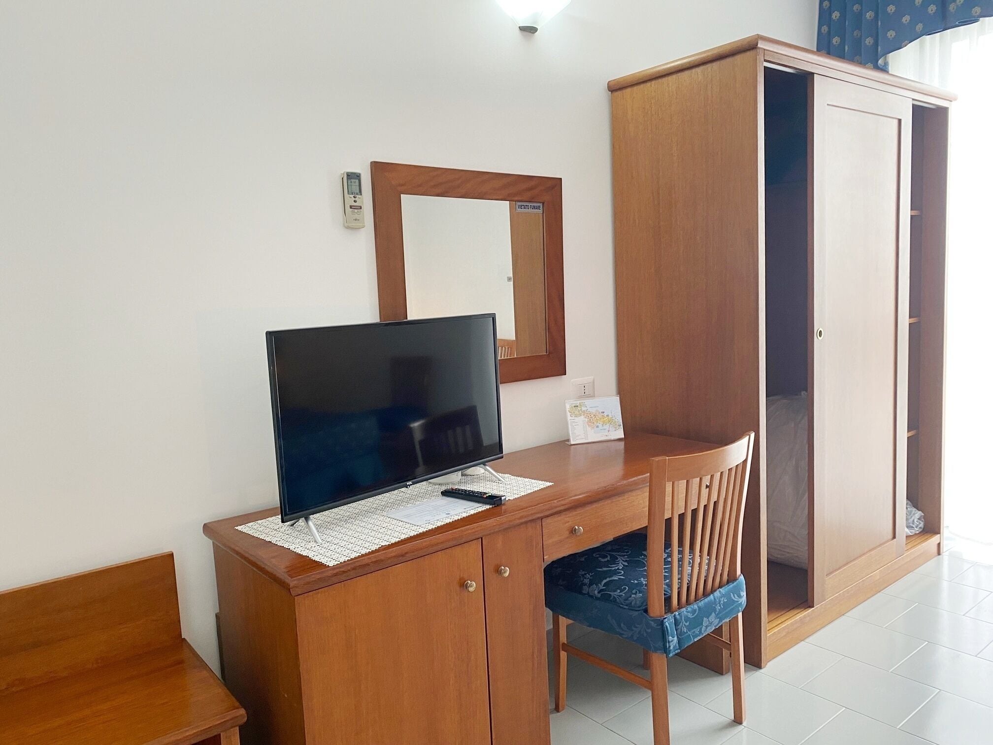 A wooden desk with a flat-screen TV, a chair, and a wardrobe in a room with white walls.