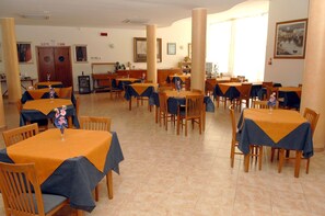 A dining area with multiple tables and chairs, some with blue tablecloths and others with orange. There are wooden chairs and a few tables with floral centerpieces.