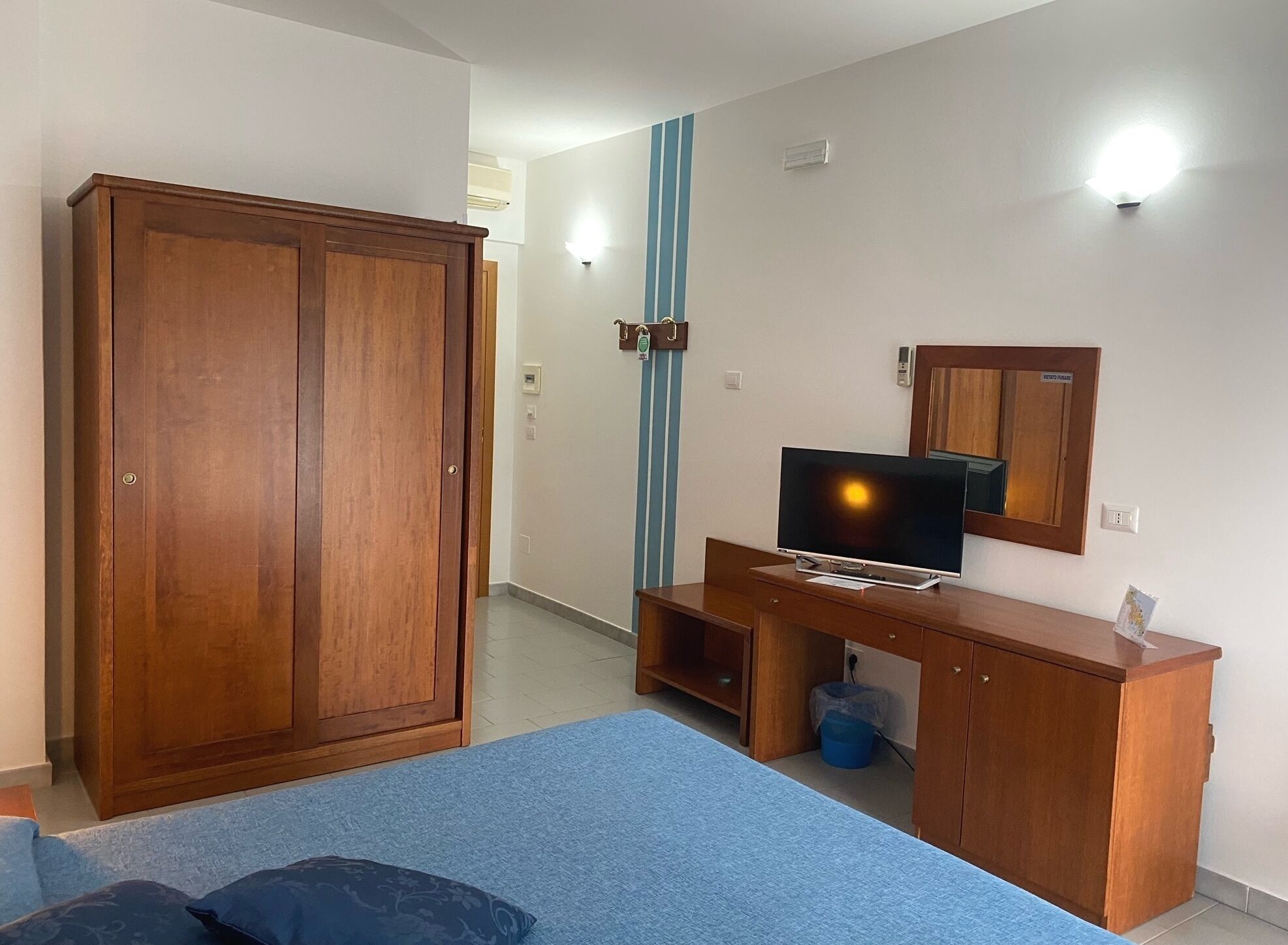 A bedroom with a bed, a wooden wardrobe, a desk with a television, and a mirror.