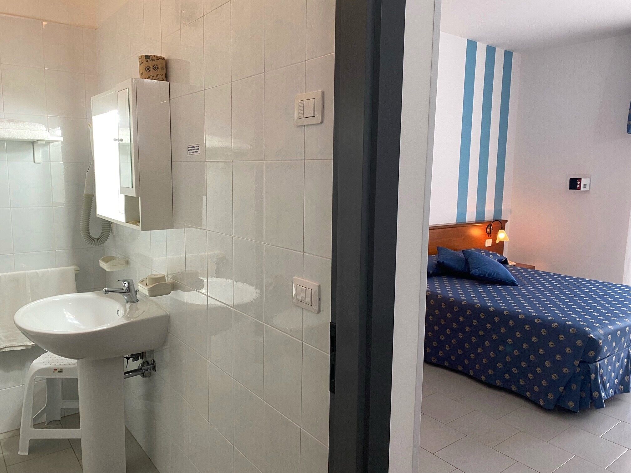A bathroom with a sink, mirror, and toilet. There is a bed with a blue bedspread and pillows in the adjacent room.
