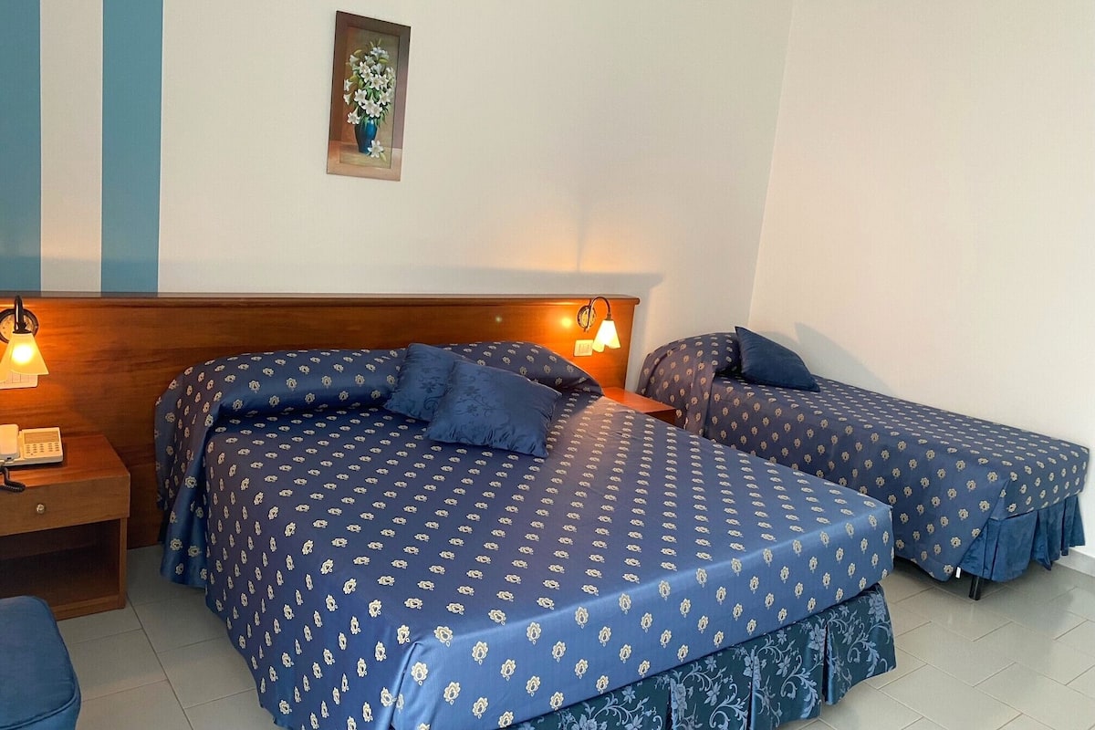 Une chambre d’hôtel avec deux lits, chacun recouvert d’une couvre-lit à motifs bleus et agrémenté d’oreillers assortis. Un petit tableau est accroché au mur au-dessus des lits, une table de chevet avec une lampe s’y trouve, ainsi qu’un téléphone sur la table de nuit.
