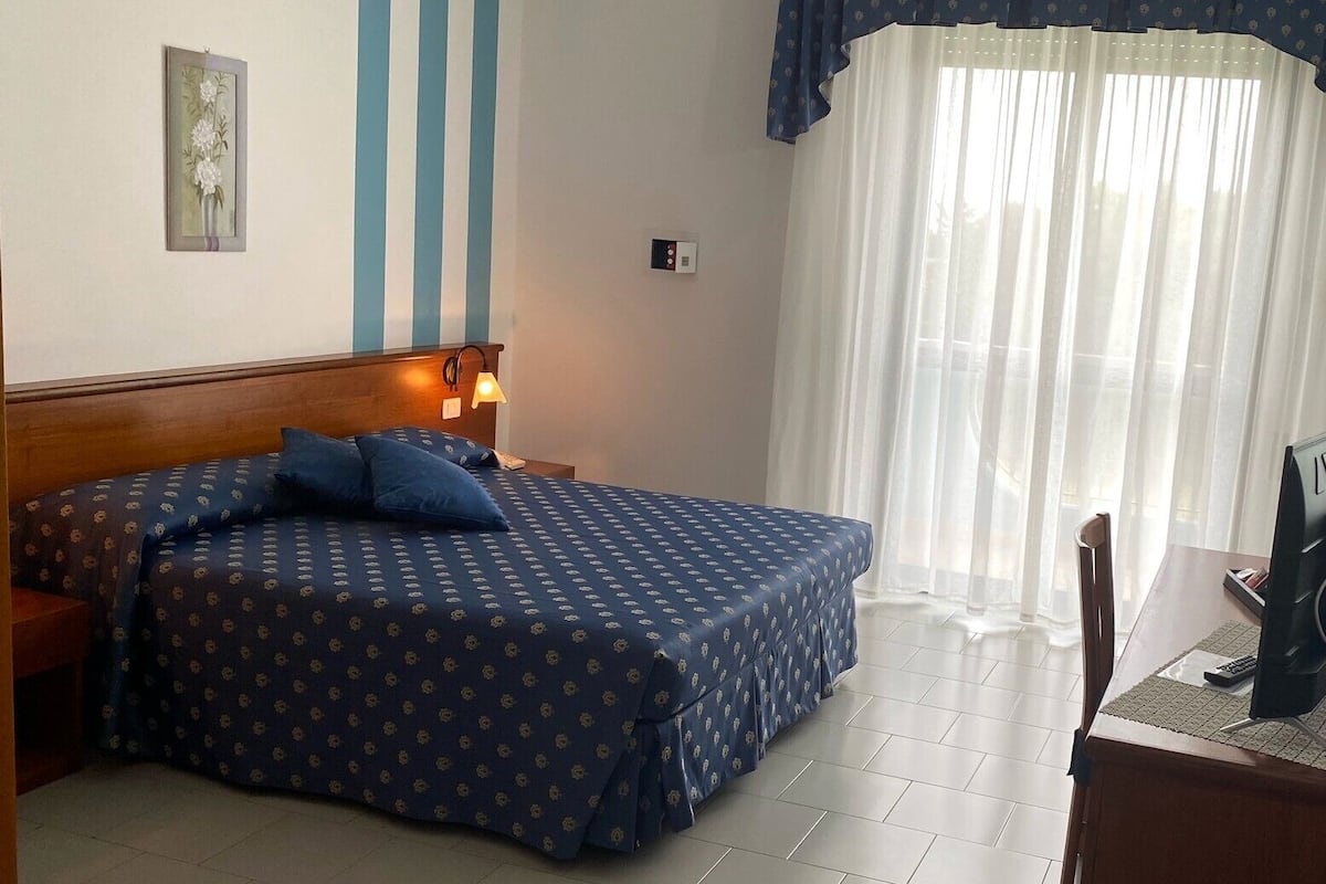 Une chambre d’hôtel avec un lit, un bureau, une chaise, une lampe et une fenêtre avec des rideaux.