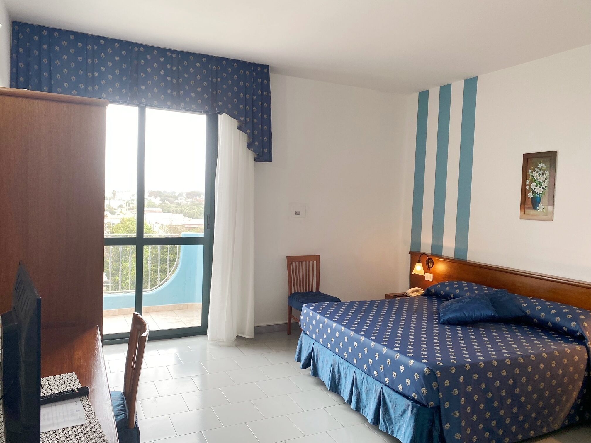 A hotel room with a bed, a desk, a chair, a small table, a lamp, and a window with curtains.