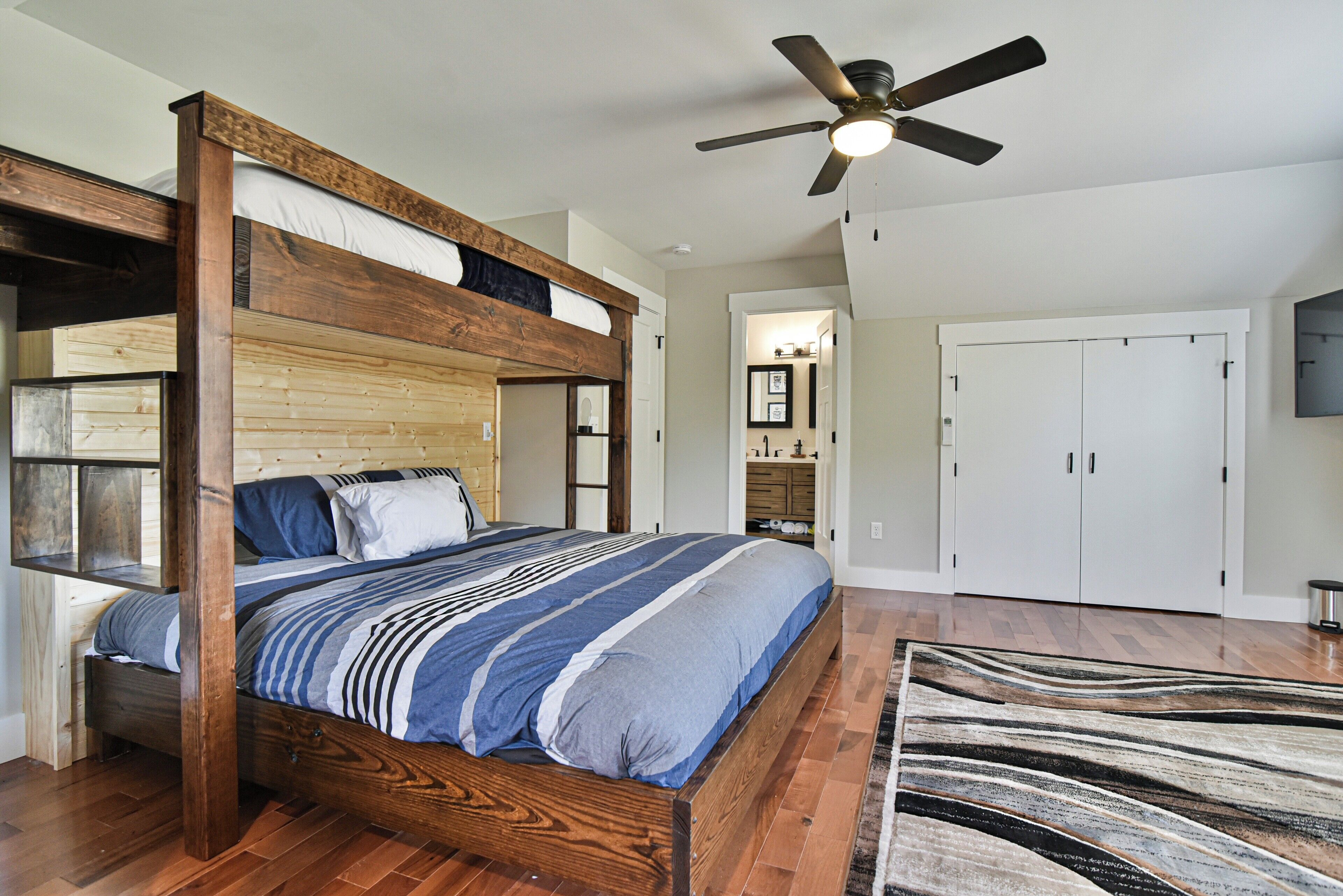 Quarto com cama beliche, ventilador de teto, piso de madeira e um tapete grande.