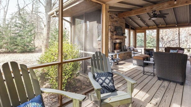 A covered outdoor area with wooden deck, chairs, a fireplace, and a view of trees.