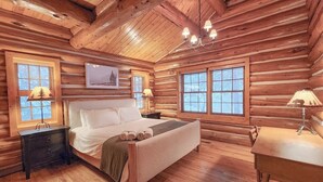 Quarto de cabana de tronco com cama, mesas de cabeceira, escrivaninha e lustre.