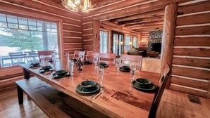 Área de jantar de uma cabana rústica de madeira, com uma mesa comprida posta para refeição, bancos e uma lareira ao fundo.